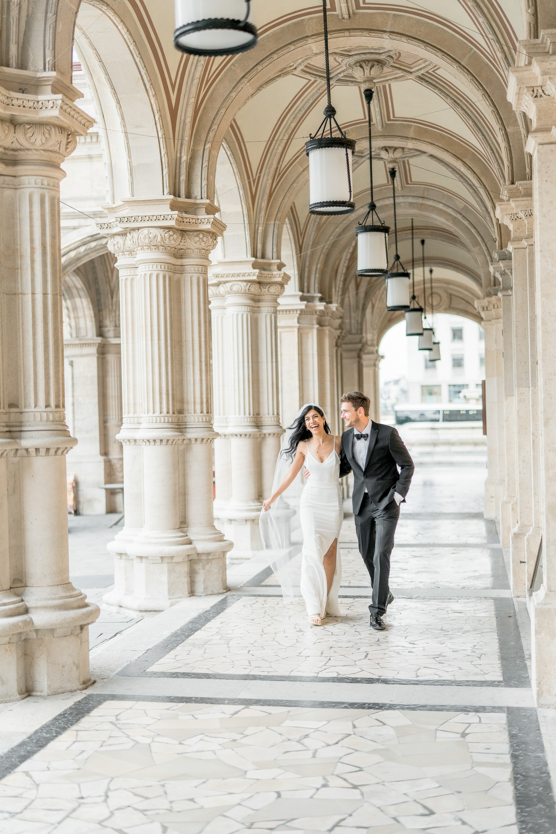 Hochzeitsfotograf Wien: Ein lachendes Brautpaar spaziert Arm in Arm durch einen Arkadengang der Oper.