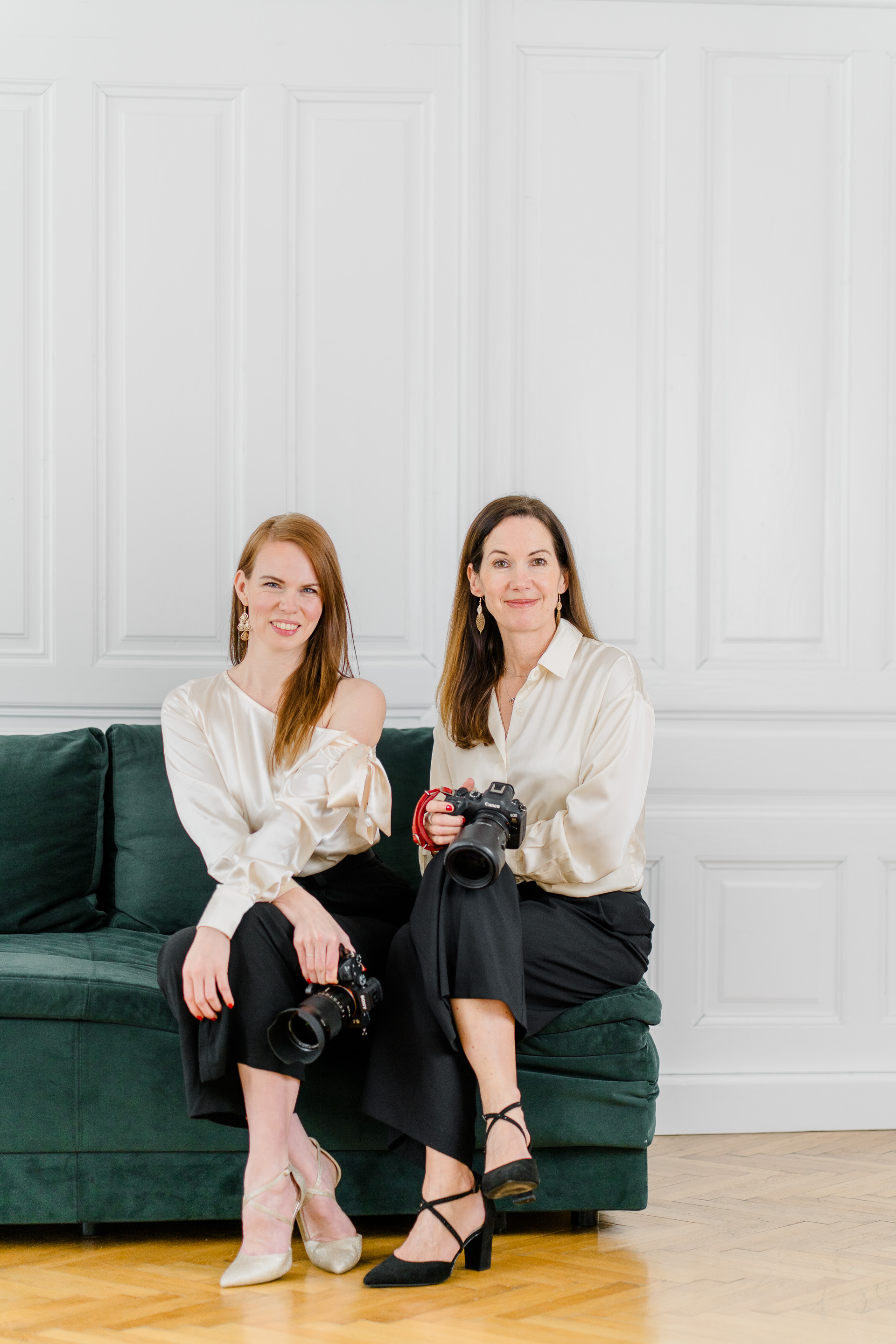 Hochzeitsfotograf Wien: Zwei Fotografinnen sitzen auf einem dunkelgrünen Sofa und lächeln in die Kamera.