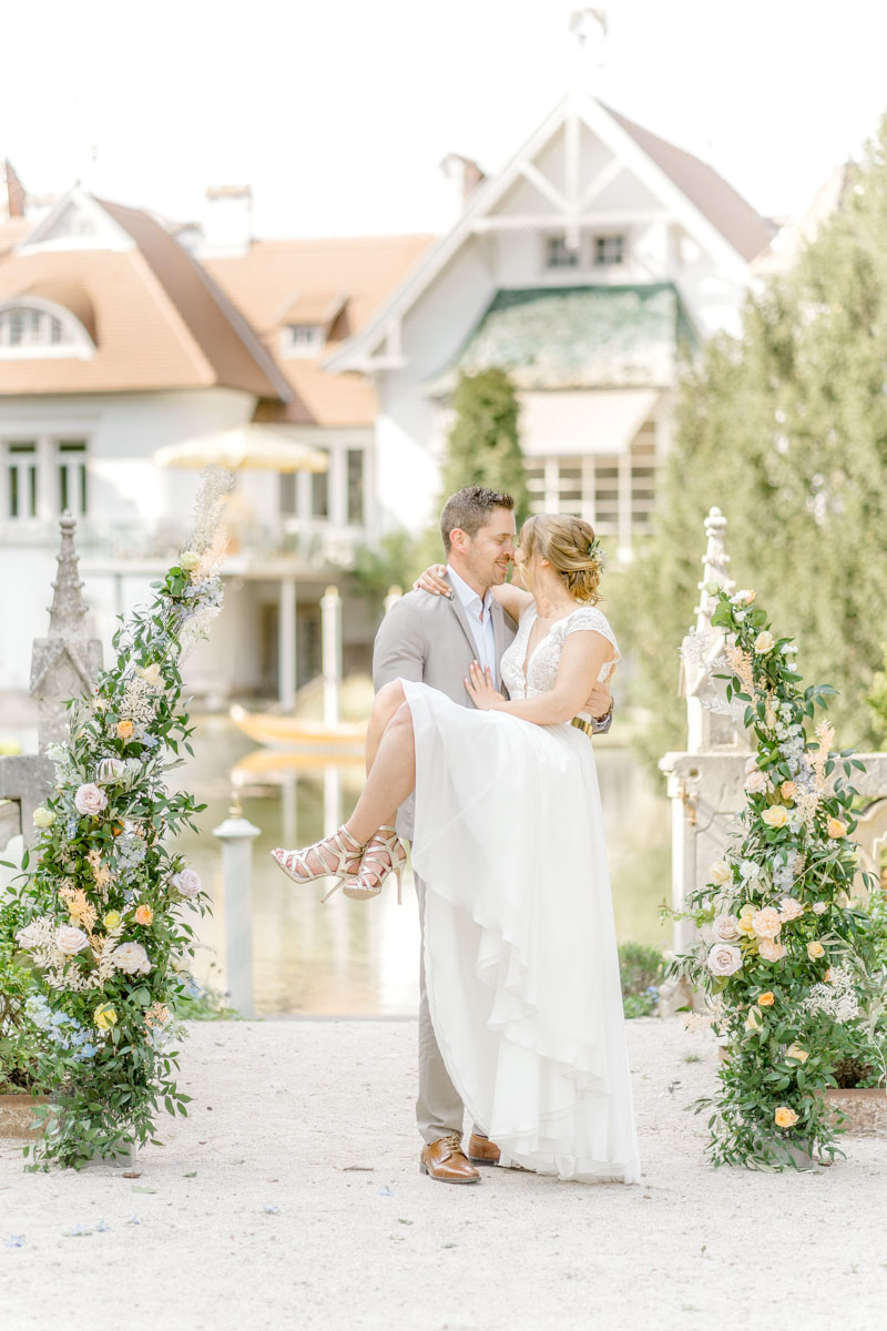 Hochzeitsfotograf Wien: Bräutigam trägt seine Braut vor prächtigem Schlosshintergrund eingerahmt von bunten Blumen.